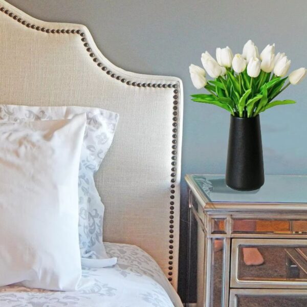 Buquê de Tulipas Artificiais Brancas em vaso preto decorando mesa de cabeceira ao lado de cama com cabeceira estofada, criando um ambiente sofisticado no quarto.