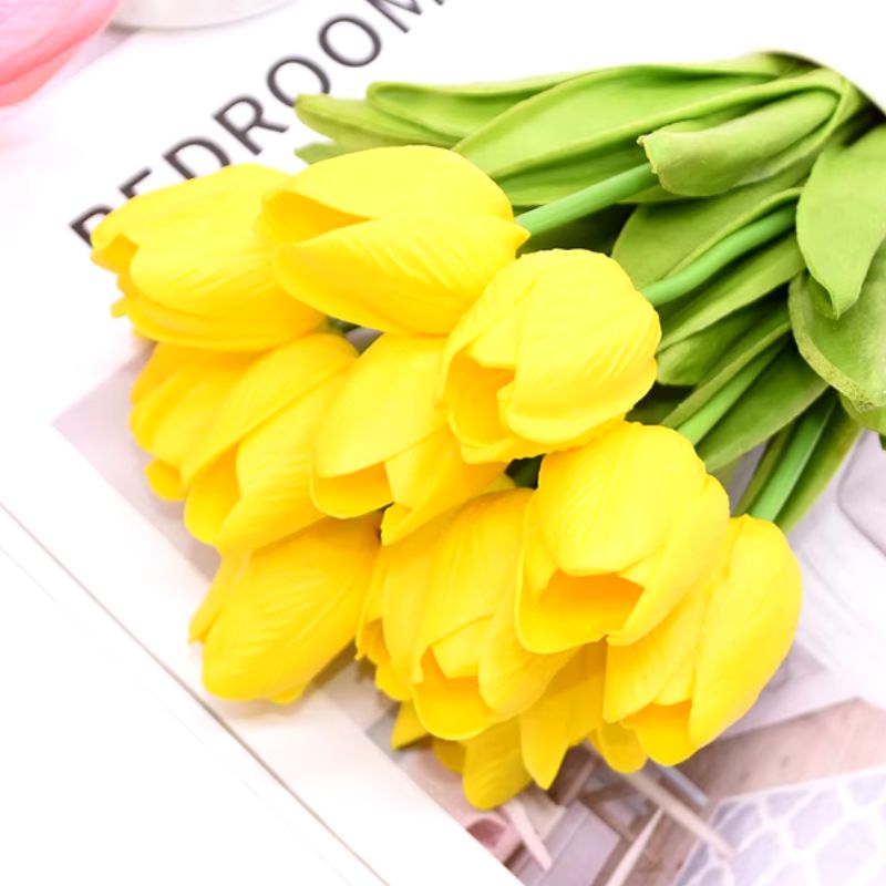 Buquê de tulipas artificiais amarelas sobre uma mesa com fundo de revista, ideal para decorar salas de estar e quartos.