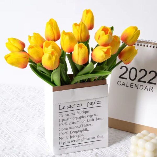 Arranjo de tulipas artificiais amarelas com bordas alaranjadas em um vaso decorativo de papel, ideal para composições modernas.